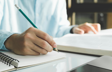 Photo of someone writing in a notebook with a pencil while referring to a book.