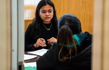 Woman takes notes as another woman speaks to her inside an office.